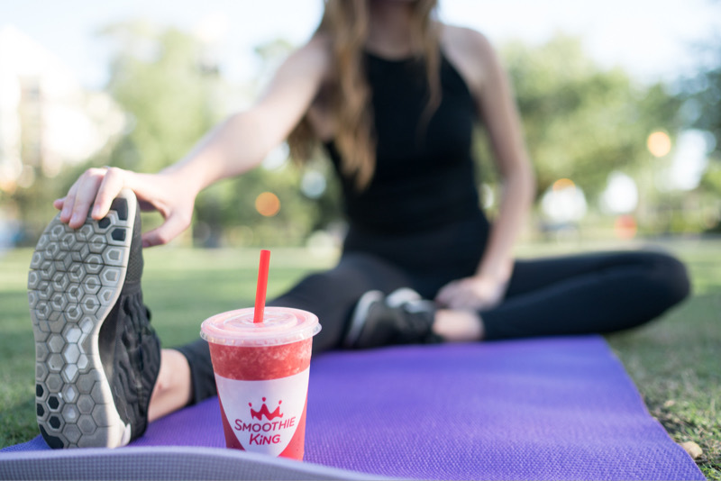 A woman stretching on a yoga mat outside with a post-workout smoothie from Smoothie King