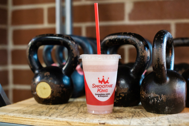Smoothie King whey protein shake sitting on a table with kettlebells behind it