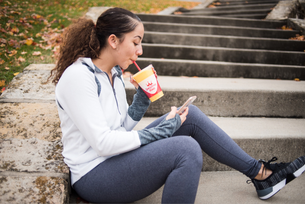 Woman drinking a Smoothie King protein smoothie and scrolling through her phone on the stairs outside after a run