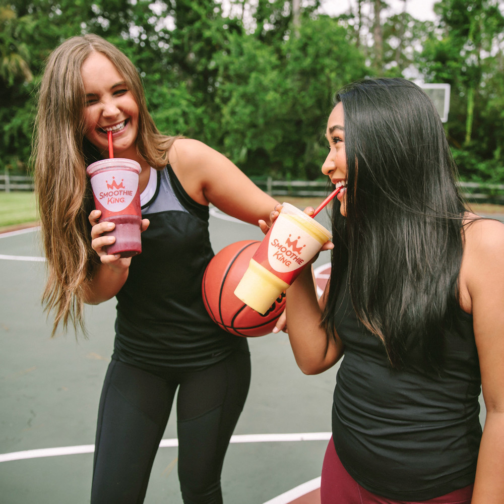 Two women hold best fruit smoothies from Smoothie King on a basketball court